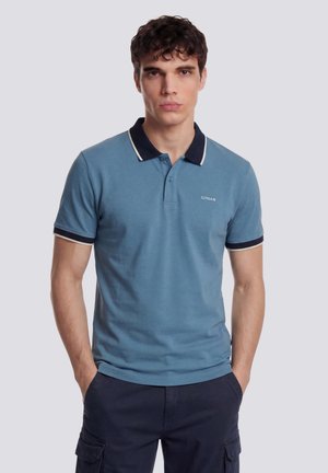 Junger Mann trägt ein blaues Polohemd mit marineblauem Besatz und dunklen Cargohosen, steht mit den Händen in den Taschen vor einem schlichten Hintergrund.