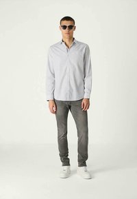 Homem com óculos de sol redondos pretos, camisa branca com padrão de botões, jeans cinzentos justos e ténis brancos, de pé contra um fundo claro liso.