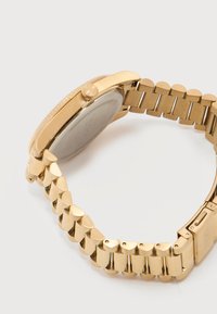 Ρολόι χειρός από μέταλλο σε χρυσό τόνο με συνδεδεμένο μπρασελέ και κλειστό κούμπωμα, εμφανίζεται πλάγια με έμφαση στο μπρασελέ και στην πίσω όψη της κάσας.