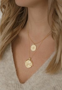 Collane d'oro a strati con un pendente circolare decorato da motivi intricati e un disco più piccolo con una stella impressa, posate su un morbido maglione grigio.