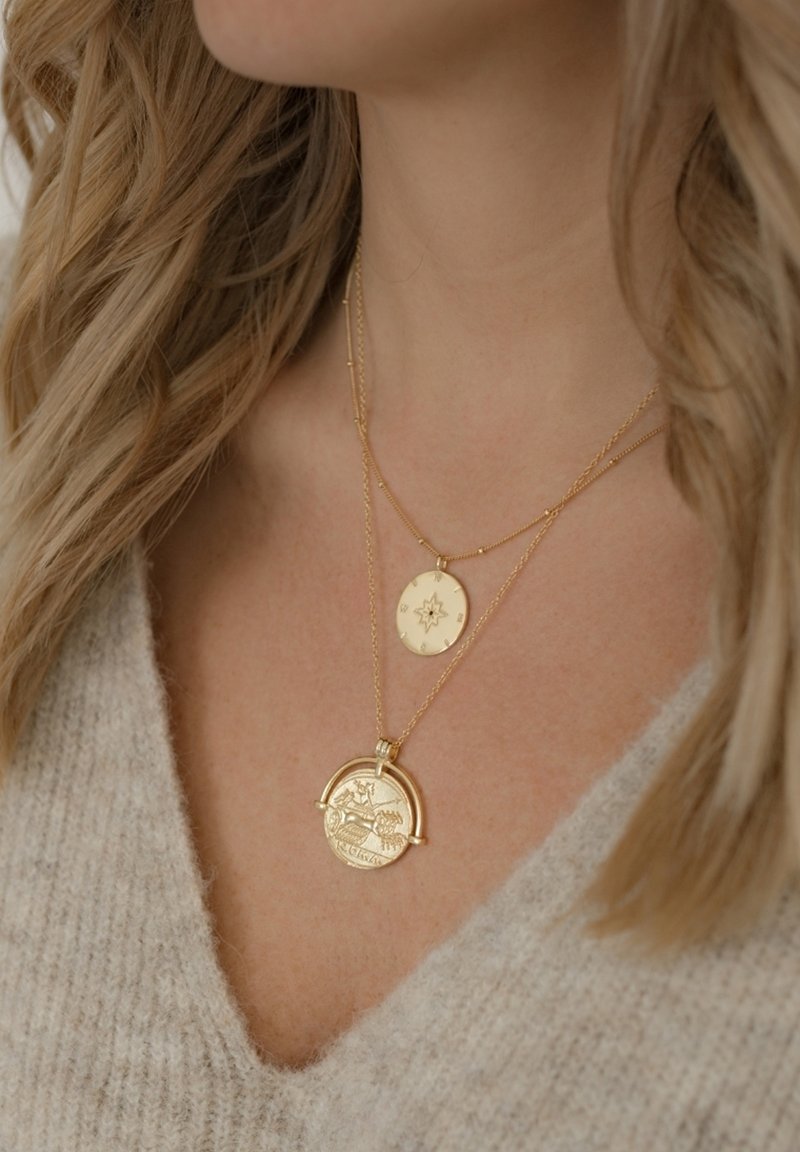 Collane d'oro a strati con un pendente circolare decorato da motivi intricati e un disco più piccolo con una stella impressa, posate su un morbido maglione grigio.