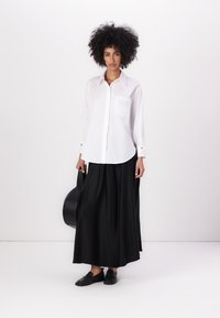 White button-up shirt with a pocket, paired with a flowing black maxi skirt. Dark shoes and a black handbag complement the outfit.