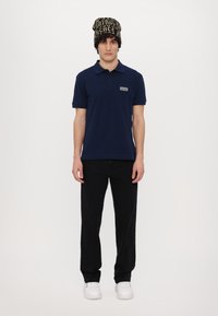 Modra polo majica s kratkimi rukavi in logotipom na prsih, v kombinaciji s črnimi hlačami ravnega kroja in belimi supergami, ki jo dopolnjuje vzorčast zimska kapa.