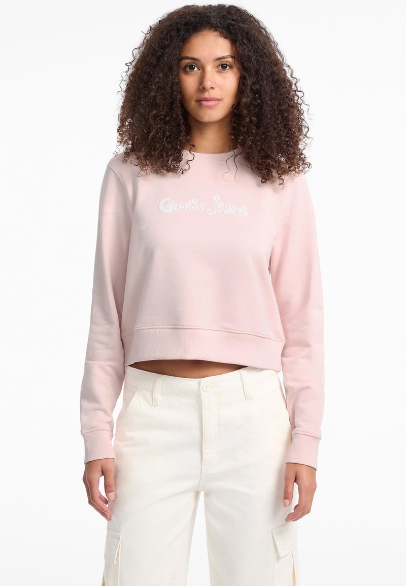 Light pink cropped sweatshirt with embroidered "Guess Jeans" logo in white, featuring long sleeves and a relaxed fit. Paired with cream-colored pants.