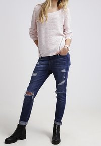 Donna che indossa un maglione a maglia rosa chiaro, jeans scuri strappati con risvolti sulle caviglie, stivali neri con zip davanti, in piedi con una mano in tasca.