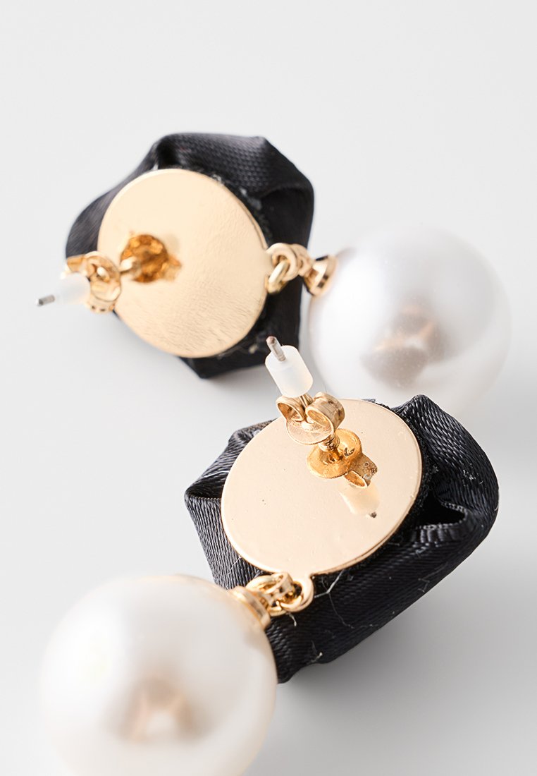 Los pendientes en tono dorado cuentan con un fondo de tela negra, grandes perlas blancas y cierres de postales seguros. Superficies suaves con acentos brillantes.