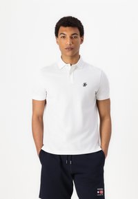 White polo shirt with a collared neck, short sleeves, and a small navy logo on the chest, paired with navy shorts.