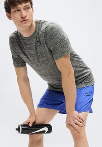 Γκρι ριγέ αθλητική μπλούζα με στρογγυλή λαιμόκοψη, σε συνδυασμό με μπλε σορτς. Κρατάει ένα μαύρο μπουκάλι νερού με λευκό λογότυπο Nike.