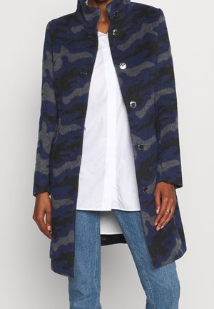 Kvinde iført en blå og grå camouflage-mønsteret jakke med sorte knapper, kombineret med en hvid skjorte og blå jeans.