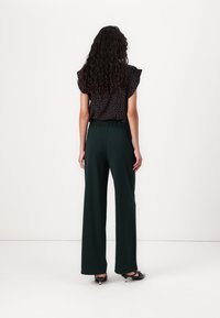 Zwarte bloemenblouse met ruches aan de mouwen, gecombineerd met hooggeknoopte, wijde groene broeken. Schoeisel bestaat uit zwarte lage hakken.