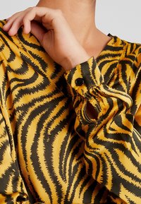 Persona che indossa una blusa a maniche lunghe a strisce tigre gialle e nere con polsini abbottonati, mano che tocca delicatamente il collo.