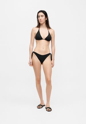 Woman standing against plain background wearing a black halter bikini with side-tie bottoms and black slide sandals.