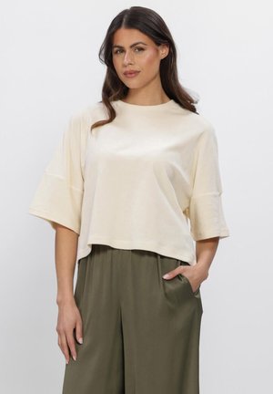 Frau mit langen dunklen Haaren, die ein locker sitzendes cremefarbenes Kurzarmtop und olivgrüne, weit geschnittene Hosen trägt, eine Hand in der Tasche.