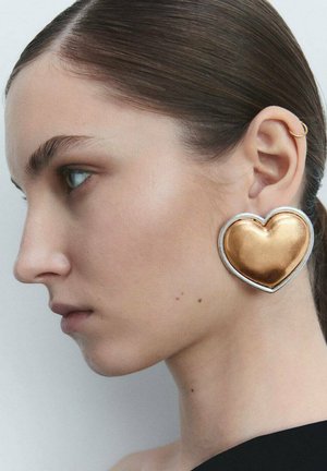 Gold heart-shaped earring with a shiny surface and a silver border, worn on a smooth, light-skinned ear against a neutral background.