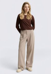 Top marrone a costine a maniche lunghe abbinato a pantaloni beige chiaro a vita alta con pieghe. La modella indossa scarpe platform beige. Sfondo semplice.