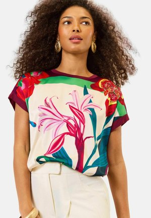 Käärmekarva juustega naine, kes kannab kõrvarõngaid ja lillemustriga särk suuremate roosade ja punaste lilledega, kokku pandud kõrge taljega valgete pükstega.