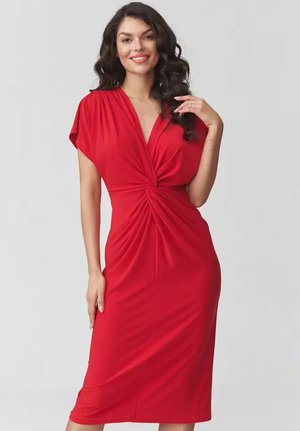 Kobieta z długimi kręconymi włosami uśmiechająca się, ubraną w czerwoną sukienkę do kolan z detalem w postaci skręconego węzła w talii.