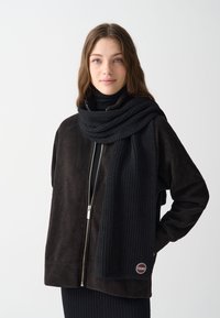 Sort strikket tørklæde draperet over en mørkebrun lynlåssløjfede jakke med ribbet tekstur, der har et rundt logo mærke i den nederste kant.