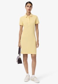 Robe polo jaune avec une coupe ajustée, des manches courtes et une patte de boutonnage. Associée à des baskets blanches et un petit sac à main violet.