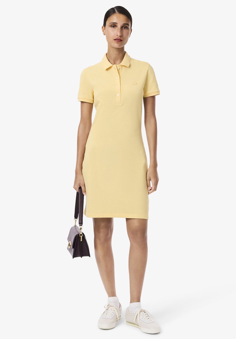 Robe polo jaune avec une coupe ajustée, des manches courtes et une patte de boutonnage. Associée à des baskets blanches et un petit sac à main violet.