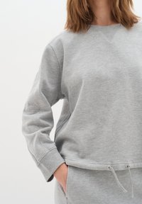 Sudadera gris con cuello redondo, puños acanalados y dobladillo fruncido con cordones. Tela suave con un acabado ligeramente texturizado.