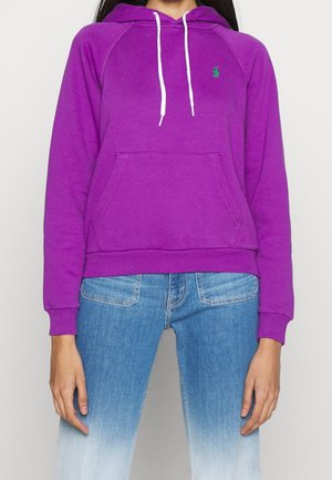 Femme portant un sweat à capuche violet avec des cordons blancs et un logo vert, associé à un jean bleu délavé.