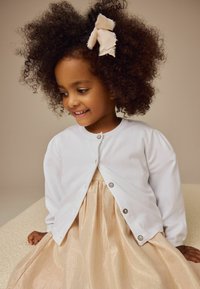 Gilet blanc avec des détails de boutons sur une robe dorée et brilliante. L'enfant a des cheveux bouclés et un ruban de couleur claire dans les cheveux.