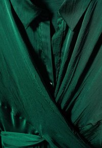 Tissu soyeux vert foncé avec un large col et un style portefeuille, montrant la texture et des plis subtils sous un éclairage doux.