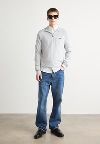 Lys grå zip-up genser over en hvit skjorte med krage, kombinert med blå jeans og svarte loafers. Modellen har på seg svarte solbriller.
