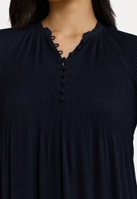 Kvinna som bär en plisserad svart blus med tygtäckta knappar och ögla stängningar längs den främre halslinjen.