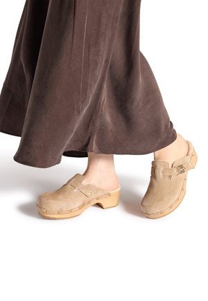 Beige semišové dřeváky s dřevěnou podrážkou, s páskem a dekorativními zlatými cvoky. Nositelka má na sobě hnědou, volnou sukni.