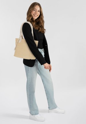 Bolso de mano tejido beige con asas de color claro, forma rectangular y textura suave. Modelo vistiendo una blusa negra y jeans de un azul claro.
