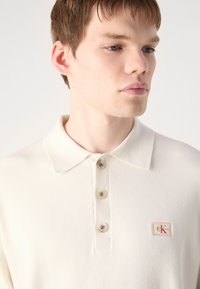 Κρέμα πόλο μπλούζα με μαλακή υφή, κλείσιμο με τρία κουμπιά και μικρό, κεντημένο λογότυπο στην αριστερή πλευρά του στήθους. Σχεδίαση κοντού γιακά.