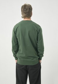 Dark green sweatshirt with long sleeves, soft texture, ribbed cuffs and hem, worn with black trousers. View from the back.