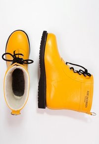 Botas de goma con cordones de color amarillo brillante y suelas negras, con forro de lana blanca, una bota en posición vertical y la otra en vista lateral mostrando la etiqueta de la marca.