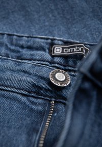 Tela de mezclilla de un azul oscuro con un botón plateado y una etiqueta negra que dice "ombri". Presenta un primer plano de las costuras y un detalle de cremallera.