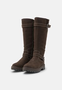 Ζευγάρι ψηλών καφέ σουέτ μποτών με χοντρές ελαστικές σόλες και διακοσμητικές ζώνες με μεταλλικές αγκράφες σε λευκό φόντο.