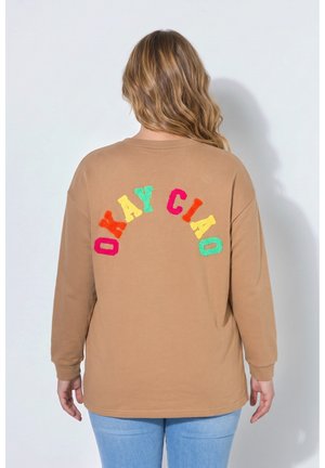 Person med langt blondt hår bærer en tanfarvet sweatshirt med farverige bogstaver, der staver "OKAY CIAO" på ryggen, og lyseblå jeans.