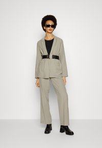 Blazer cinza claro e calças largas combinadas, usadas sobre uma blusa preta. Apresenta um cinto preto com fivela quadrada e botins pretos.