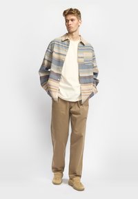 Multicolored gestreepte shirt in blauw, geel en oranje; gecombineerd met een crème t-shirt en tan wijde broek. Schoenen zijn beige.