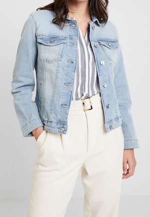 Kobieta ubrana w jasnoniebieską kurtkę dżinsową na białą i szarą koszulę w paski, zestawioną z białymi spodniami z wysokim stanem i paskiem.