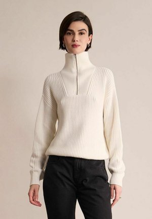 Femme aux cheveux courts et foncés porte un pull blanc côtelé avec un col montant zippé et un pantalon noir sur un fond beige uni.