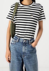 Camiseta de algodón a rayas blancas y negras con un pequeño logo gráfico en el pecho izquierdo, combinada con unos vaqueros de mezclilla azul de cintura alta.