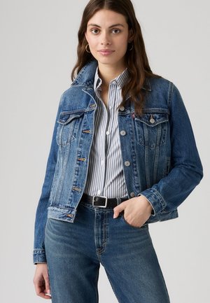 Mujer con chaqueta vaquera azul sobre una camisa de rayas con botones y vaqueros azules, con una mano en el bolsillo.