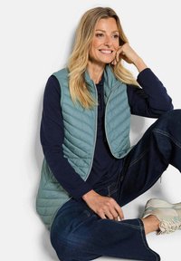 Helles türkisfarbenes Steppweste mit hohem Kragen, Reißverschluss und seitlichen Taschen, kombiniert mit einem dunkelblauen Langarmoberteil und dunklen Jeans.