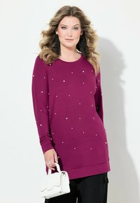Fialový top s dlouhým rukávem a kulatým výstřihem, zdobený rozptýlenými bílými ozdobami. Spárovaný s černými kalhotami a bílou kabelkou.