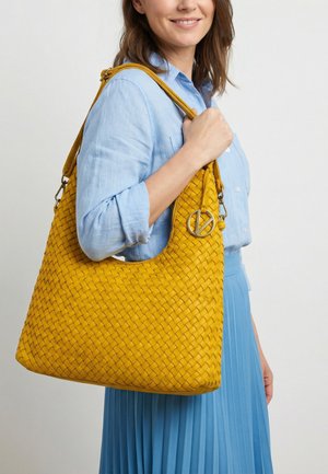 Frau trägt hellblaues Hemd und Rock, hält eine große gelbe gewebte Umhängetasche mit metallischem runden Anhänger.