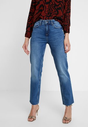 Femme portant un jean bleu taille haute à jambes droites avec des ourlets bruts, un top à manches longues rouge et noir à motifs, et des sandales à talons hauts en cuir effet serpent.