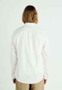 Chemise blanche à manches longues avec ourlet arrondi et poignets boutonnés, fabriquée en tissu lisse, présentée de dos sans motifs ni designs visibles.