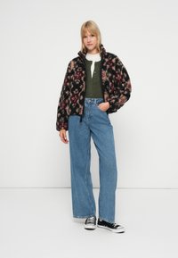 Květinová fleecová bunda v černé, růžové a krémové barvě, s vysokým límcem; v kombinaci se zelenou košilí na knoflíky a širokými modrými džíny. Černé tenisky dokončují outfit.